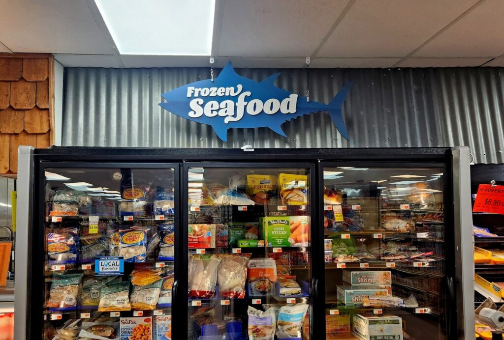 Grocery store frozen seafood section with products behind glass doors and a fish-shaped sign above.