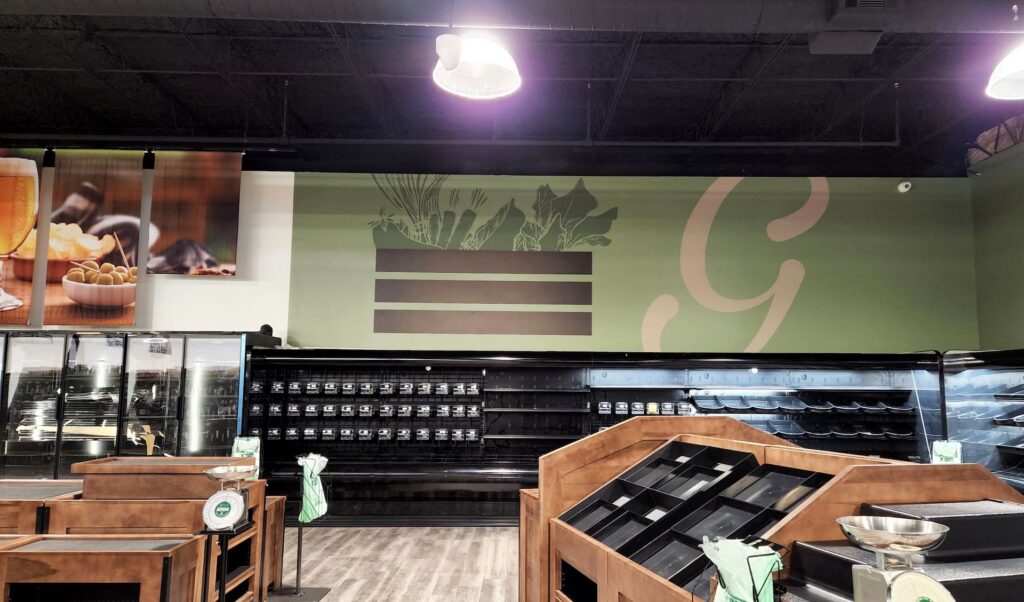 Grocery store with empty shelves and display racks, bright lighting, and a green wall featuring a vegetable graphic and large 