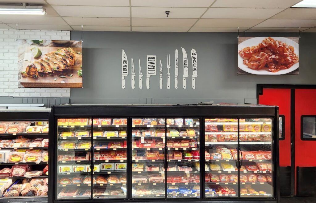 Packaged meats displayed in grocery coolers beneath decor with knife illustrations and food photos above.