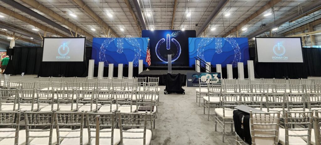 Rows of empty silver chairs face a stage with blue decor and a prominent power symbol, ready for a design-focused event.