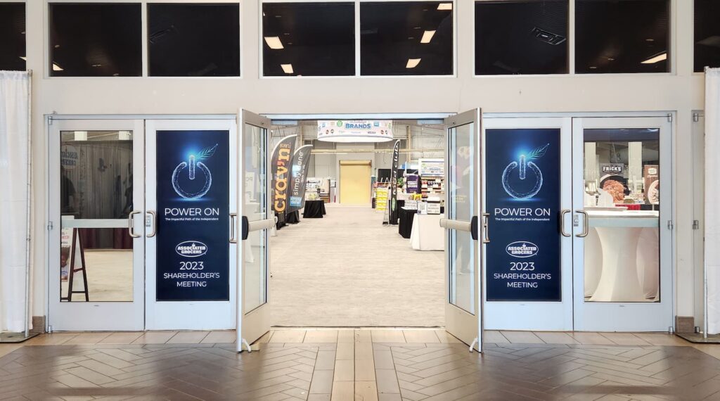Double glass doors show 2023 Shareholder’s Meeting signs; inside, Design Services booths line the convention hall.