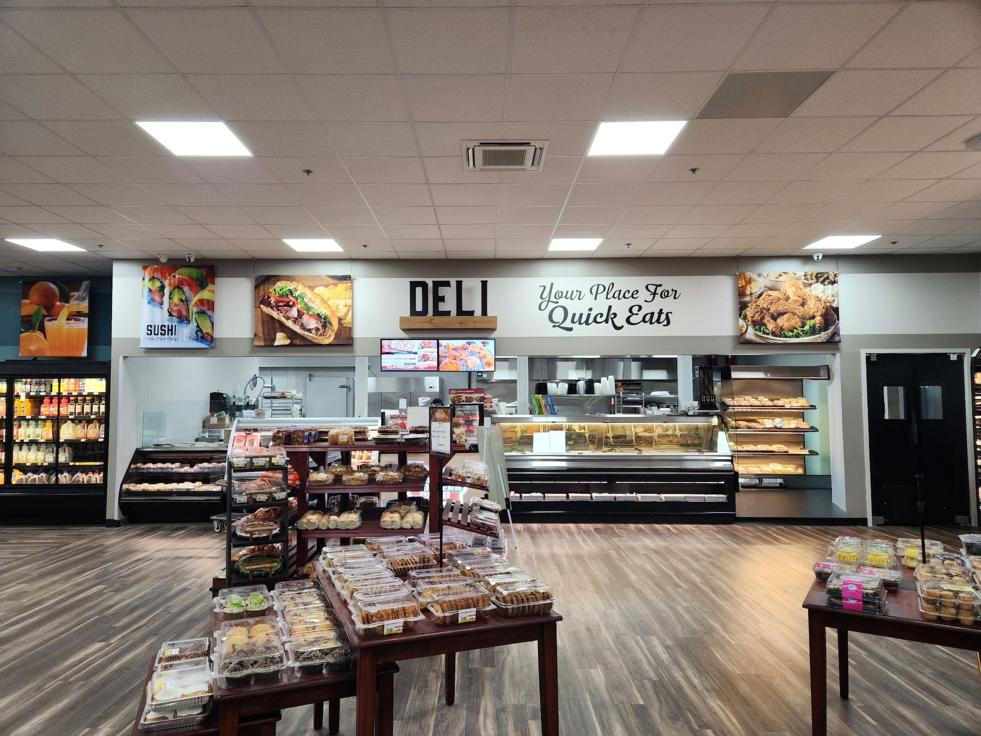 A grocery store deli counter displays prepared foods and baked goods beneath a "Your Place For Quick Eats" sign.