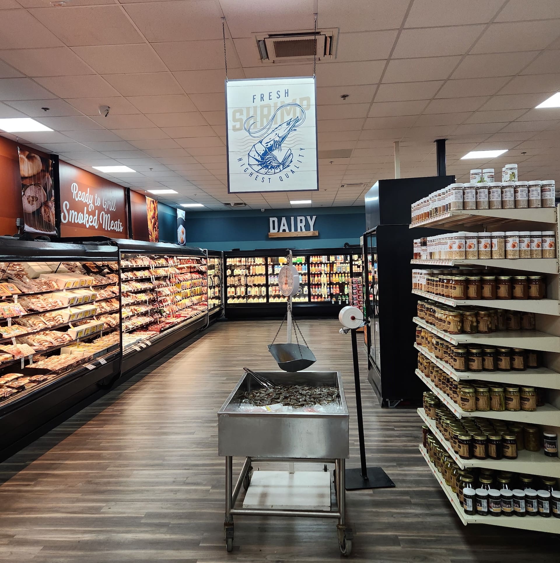 A grocery store aisle with modern décor displays seafood, canned goods, and coolers stocked with smoked meats and dairy.