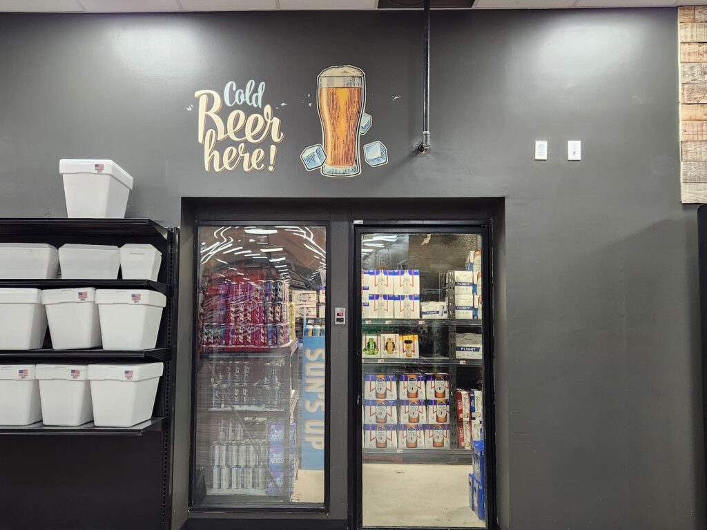A glass-door walk-in cooler displays beer boxes, with a colorful sign above that reads “Cold Beer here!” and lively retail décor.
