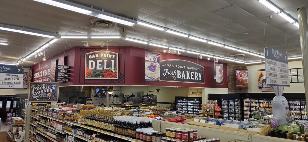 A grocery store interior with deli and bakery sections above aisles filled with packaged foods and condiments.