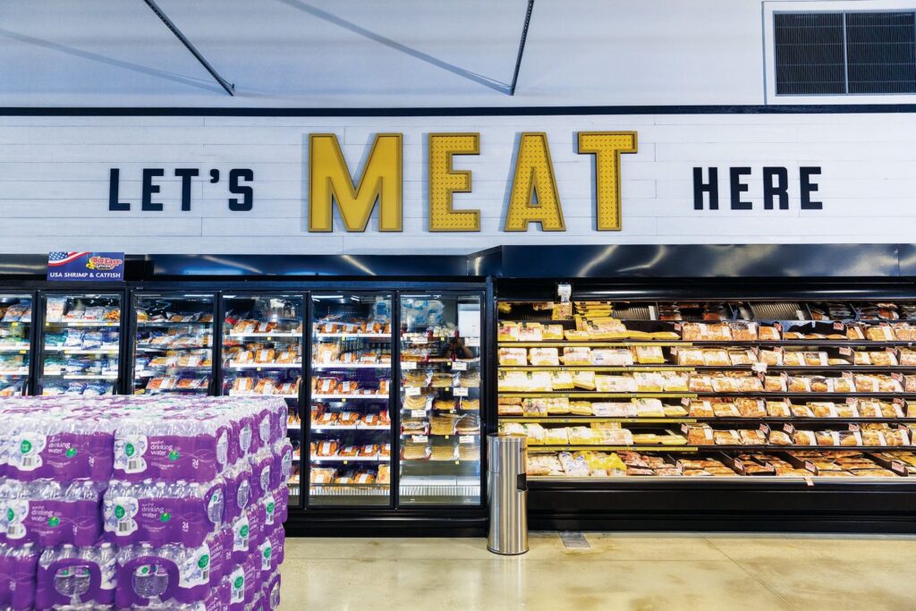 A grocery store meat section with bold decor and a “LET’S MEAT HERE” sign above shelves of neatly arranged packaged meats.