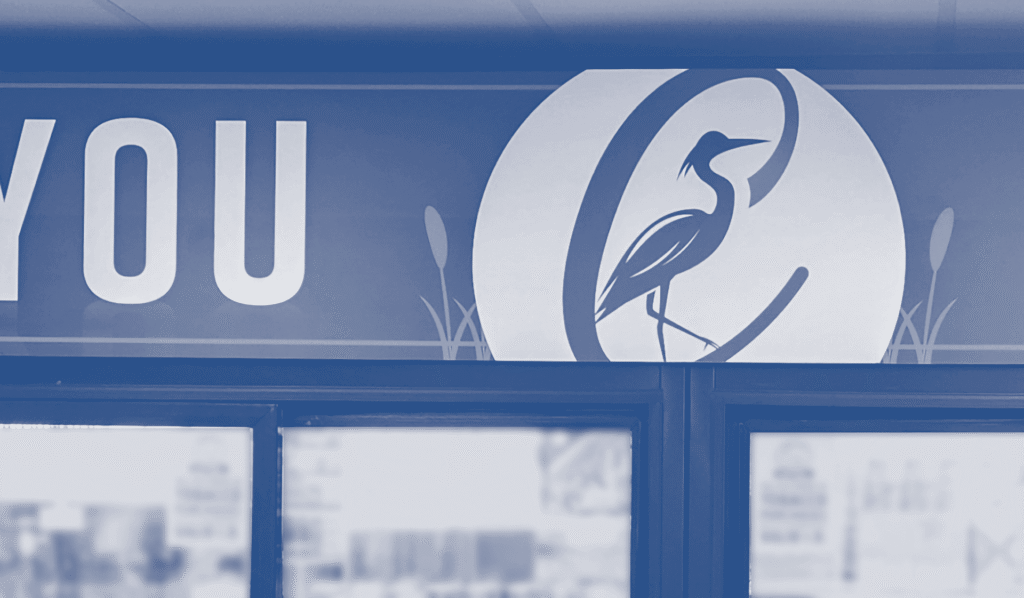 A sign displays the word "YOU" next to a logo of a bird standing before a circle with reed-like shapes.