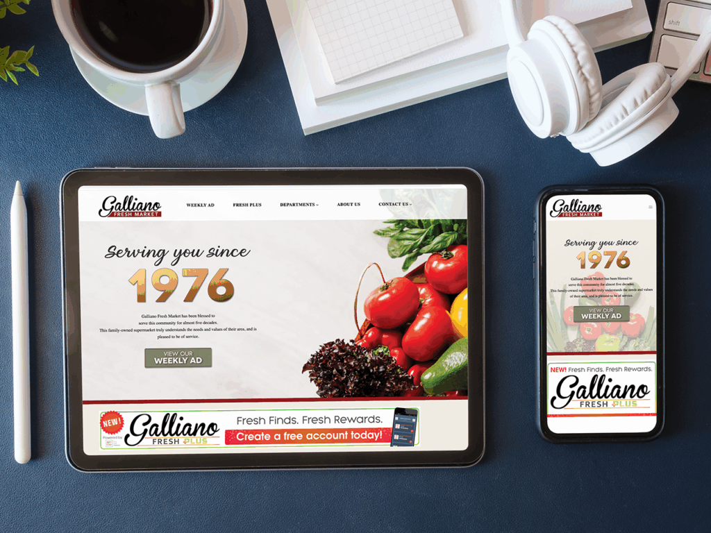 A tablet and smartphone display the Galliano Fresh Market website with vegetables, a stylus, coffee cup, notebook, and headphones nearby.