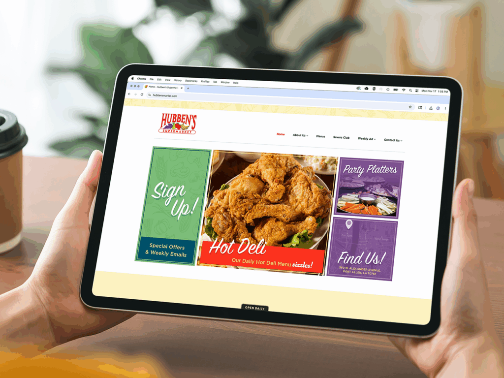 A person holds a tablet displaying a website for Hubben's Supermarket, featuring a hot deli section and sign-up and party platter options.