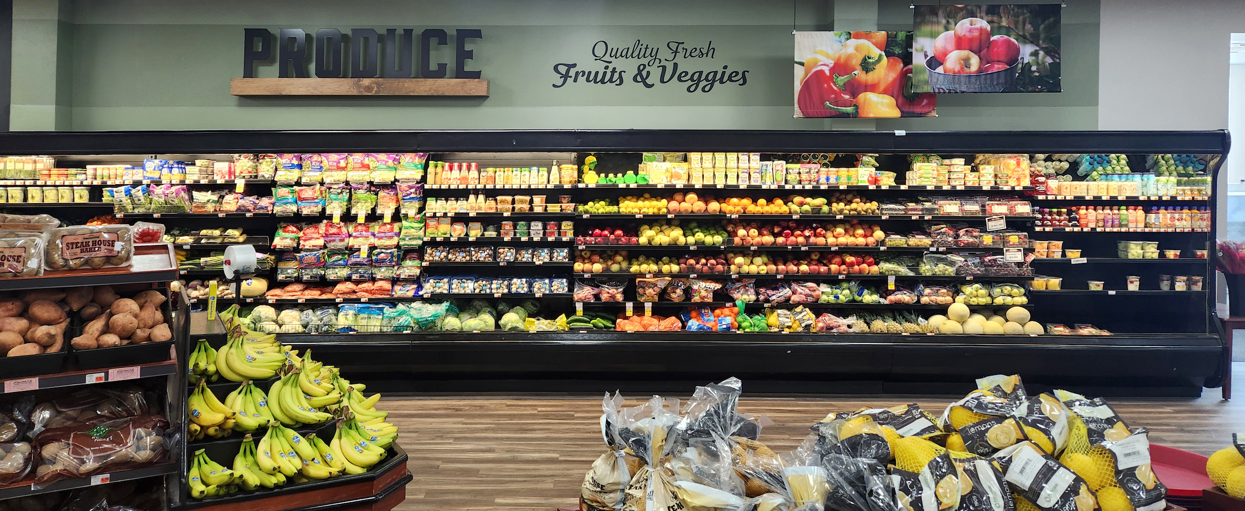 Grocery store produce section with shelves of fruits, vegetables, packaged goods, and displays of bananas and melons in the foreground.