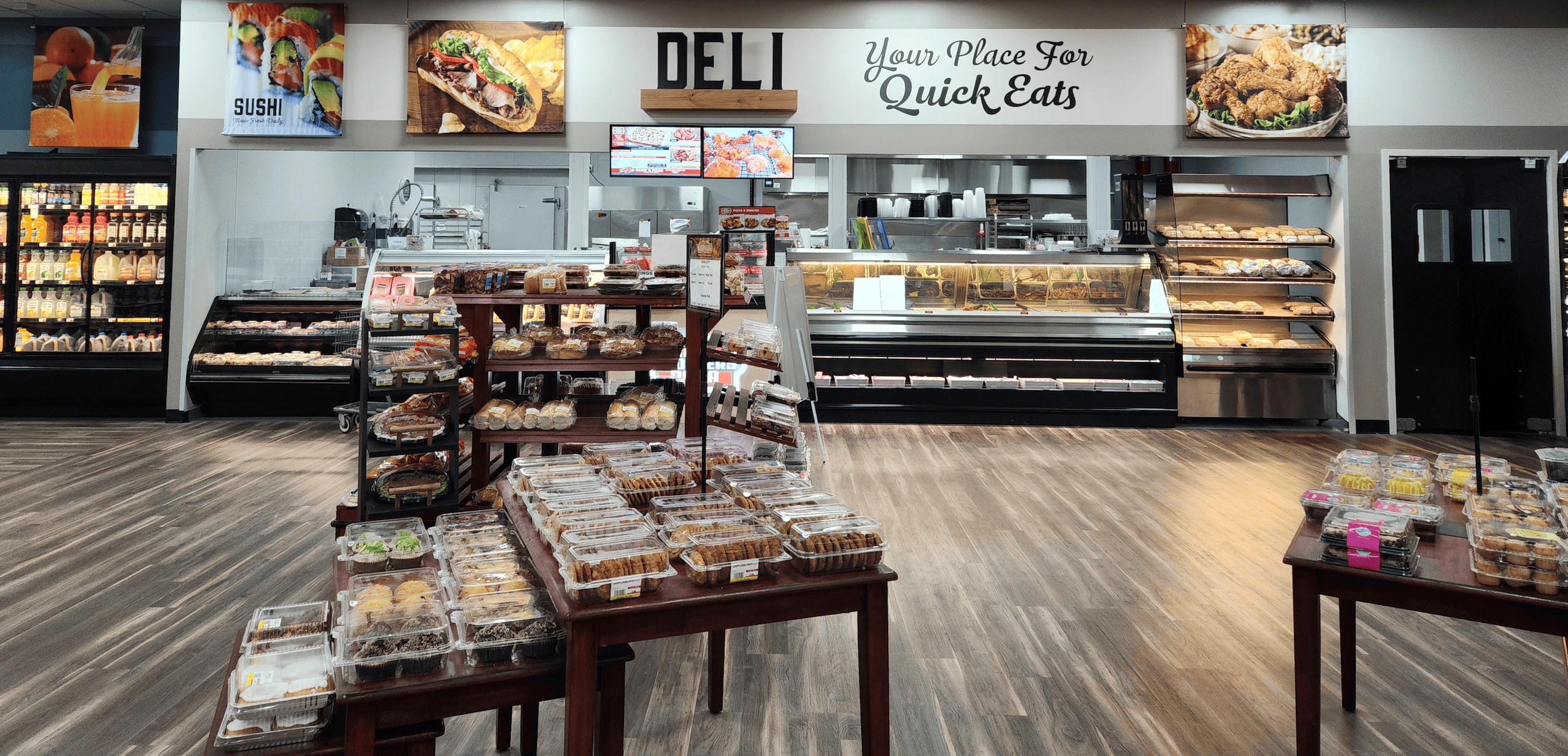A grocery store deli section with packaged baked goods on tables and shelves, and a counter displaying hot food in the background.