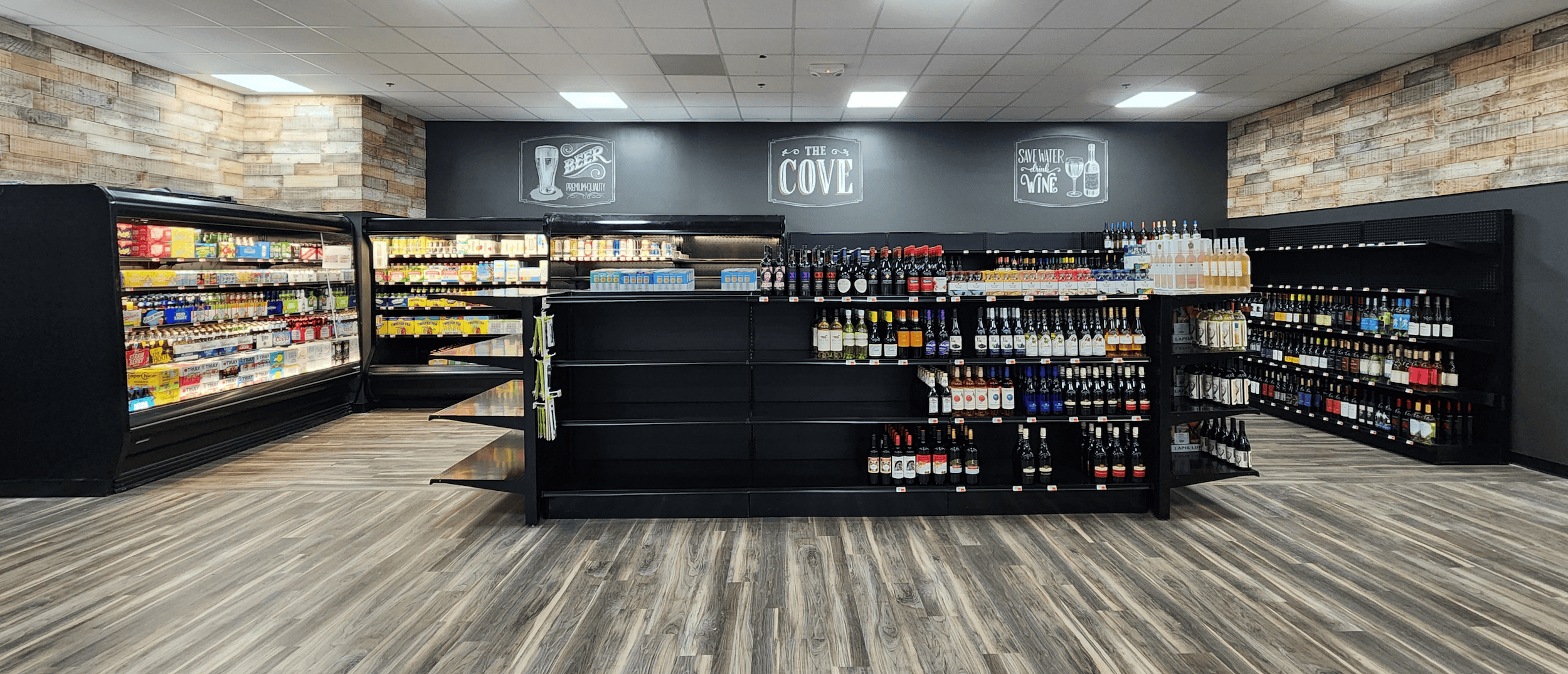 A grocery store aisle with shelves and coolers stocked with beverages, including wine, beer, and soft drinks, under bright lighting.
