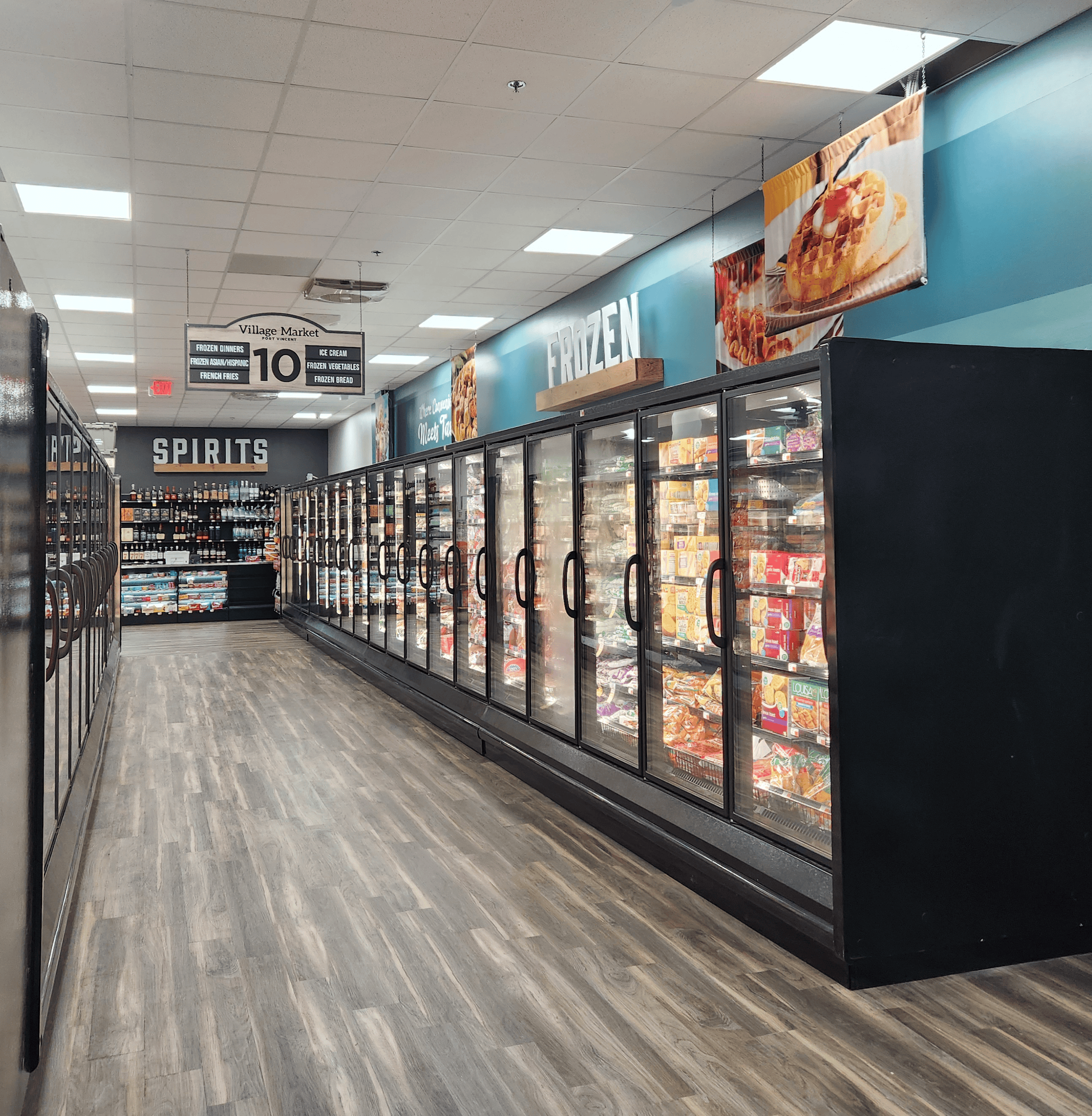 A grocery store aisle with freezers full of frozen food on the right, spirits section in the back, and wood-look flooring.