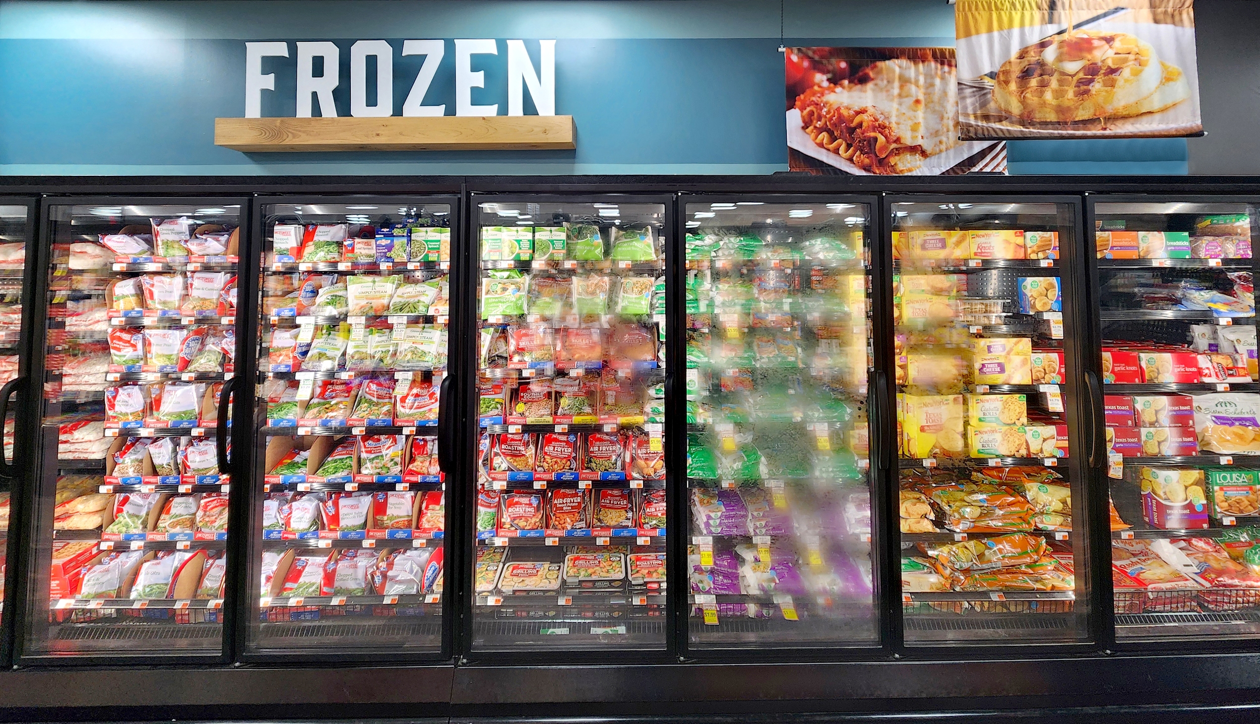 Grocery store freezer section with glass doors displaying various frozen food products and a large "FROZEN" sign above.